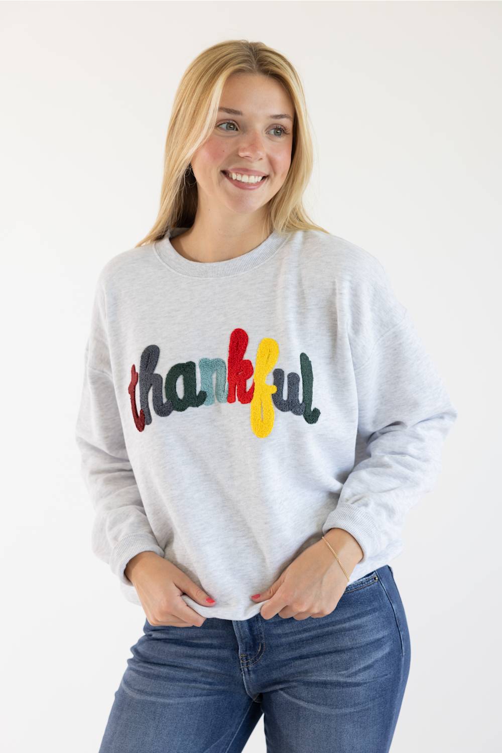 A smiling blonde woman wearing a light heather gray sweatshirt with the word 'thankful' written across the chest in colorful letters, paired with blue jeans, against a plain white background.