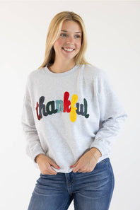 A smiling blonde woman wearing a light heather gray sweatshirt with the word 'thankful' written across the chest in colorful letters, paired with blue jeans, against a plain white background.