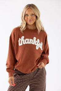 A woman wearing a rust-brown crewneck sweatshirt with 'thankful' written across the chest in white script, paired with brown and white striped pants, against a plain white background.