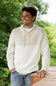A man wearing a cream-colored knit pullover hoodie with a textured chest panel and drawstrings, paired with blue jeans, standing outdoors on a wooden deck with a blurred green garden background.