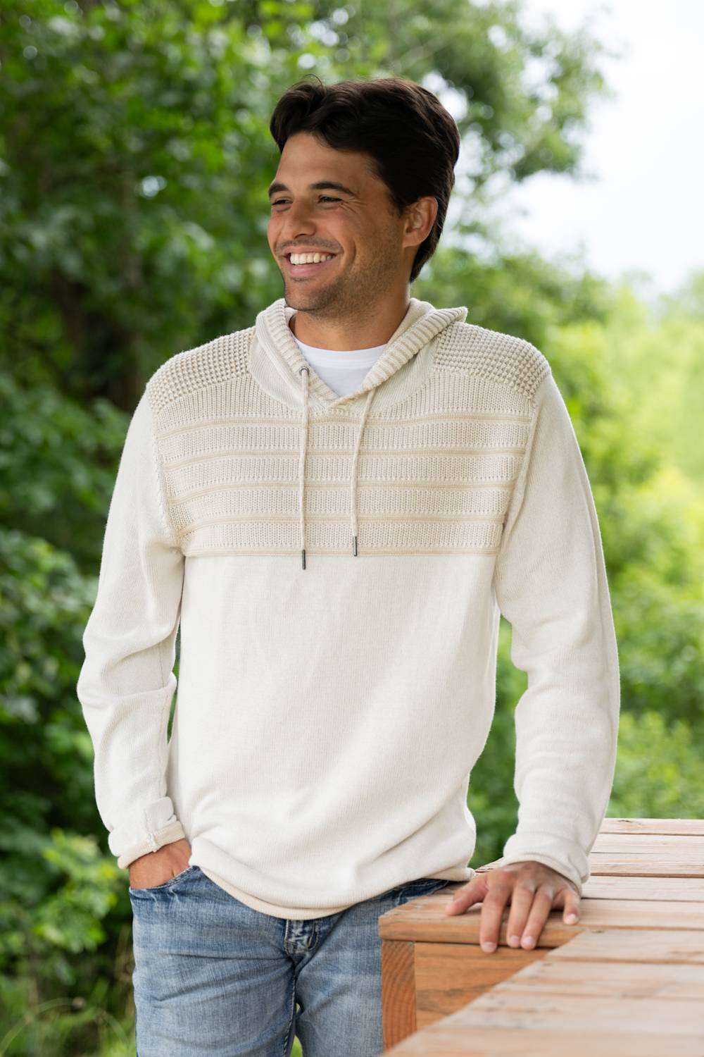 A man wearing a cream-colored knit pullover hoodie with a textured chest panel and drawstrings, paired with blue jeans, standing outdoors on a wooden deck with a blurred green garden background.