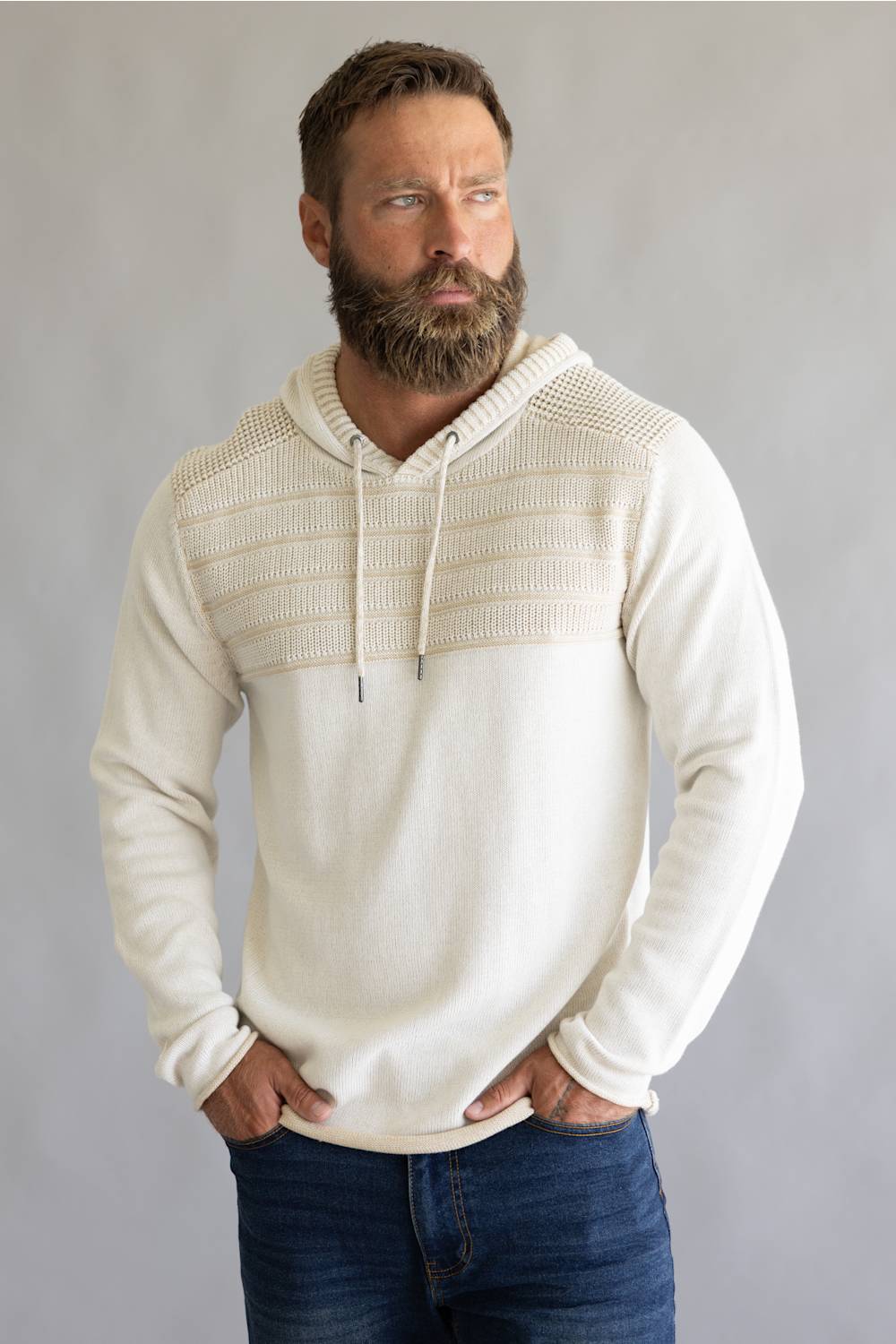 A bearded man wearing a cream-colored knit hoodie with a textured, ribbed yoke across the chest, paired with blue jeans, standing against a neutral gray studio background.