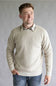A smiling man wearing a light beige ribbed knit crewneck sweater over a plaid collared shirt and blue jeans, standing against a neutral gray studio background.