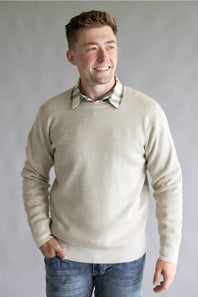 A smiling man wearing a light beige ribbed knit crewneck sweater over a plaid collared shirt and blue jeans, standing against a neutral gray studio background.