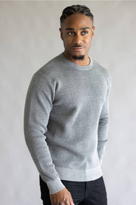 A man wearing a light gray crewneck knit sweater and dark trousers, standing against a neutral light gray background.