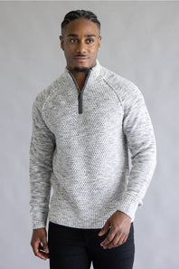 A man wearing a light gray and white textured knit quarter-zip sweater with black pants, standing against a neutral gray studio background.