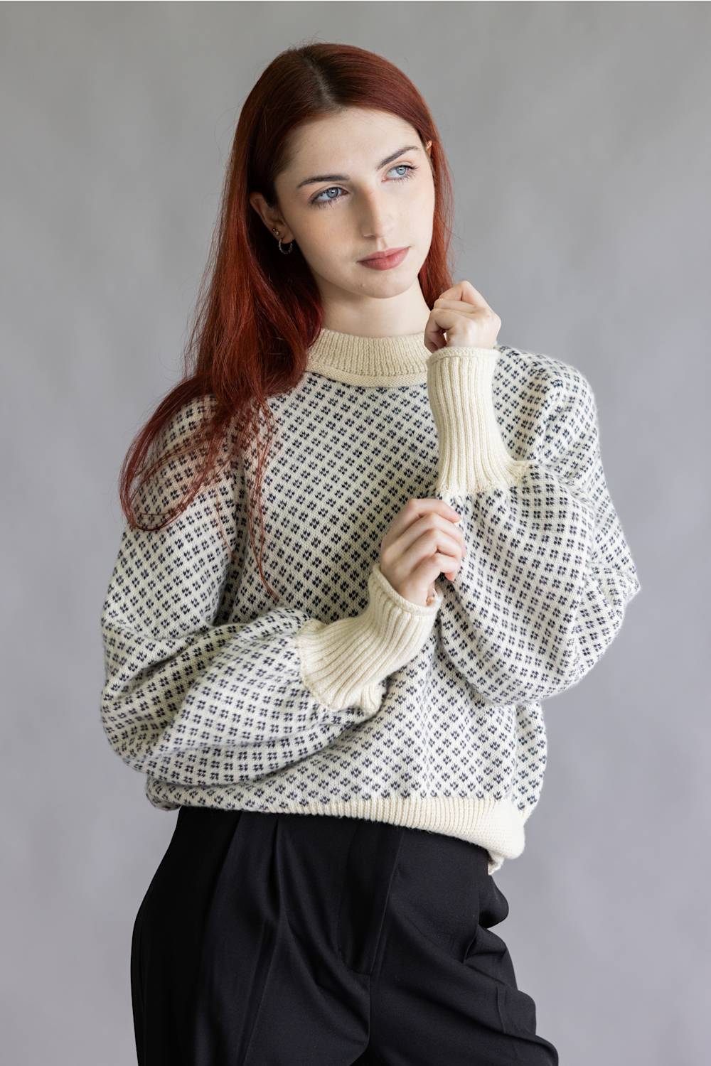 A woman with long red hair wearing a cream and black geometric patterned knit sweater with ribbed cuffs and hem, paired with black pants, against a plain gray studio background.