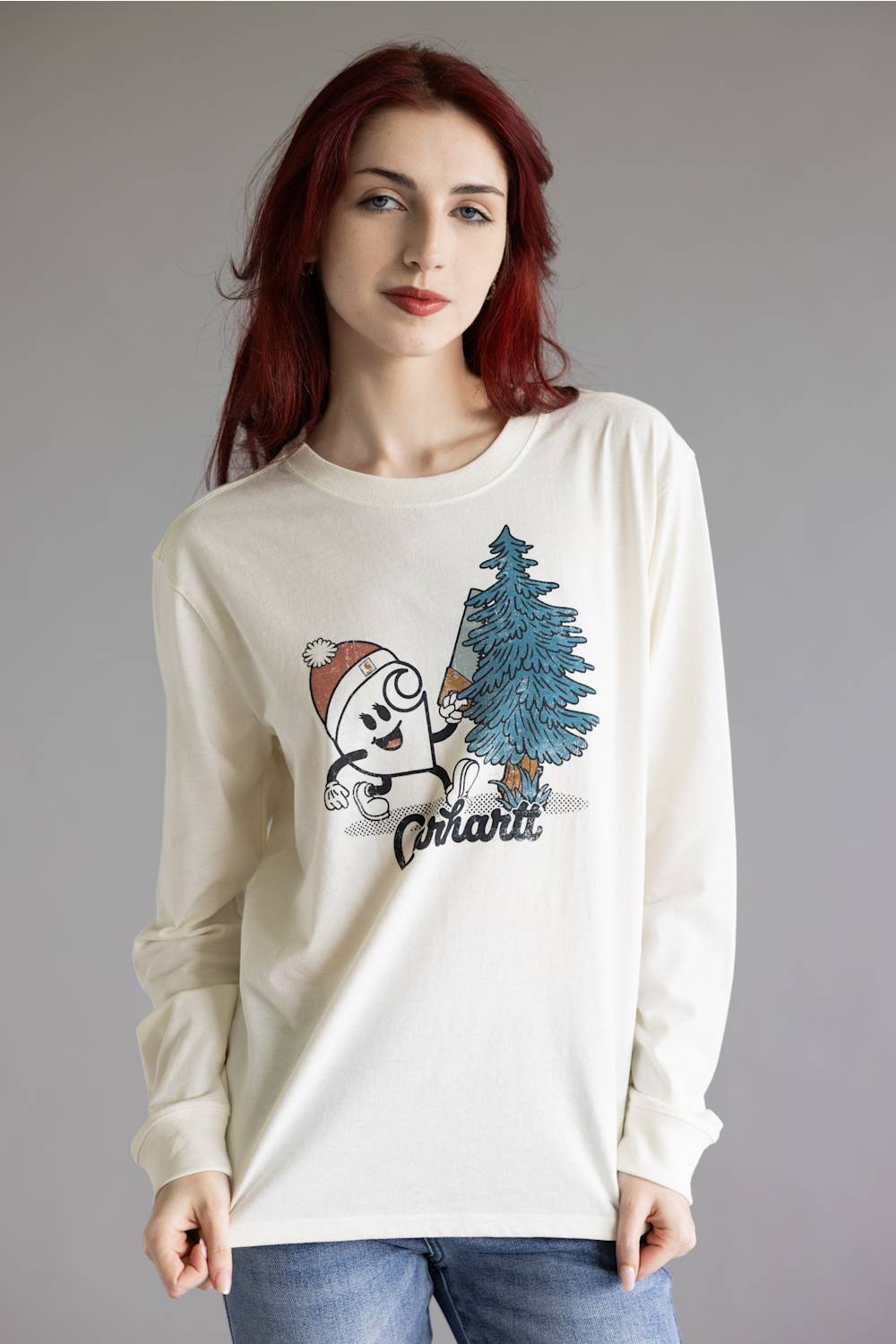 A woman with red hair wearing a cream-colored long-sleeve graphic t-shirt with a cartoon character and a blue Christmas tree, paired with blue jeans, against a plain gray studio background.