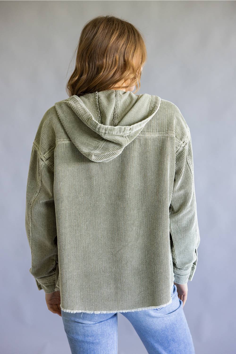A person shown from the back wearing an oversized light sage green corduroy hooded jacket over a dark top and light blue jeans, against a plain gray studio background.