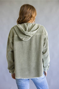A person shown from the back wearing an oversized light sage green corduroy hooded jacket over a dark top and light blue jeans, against a plain gray studio background.