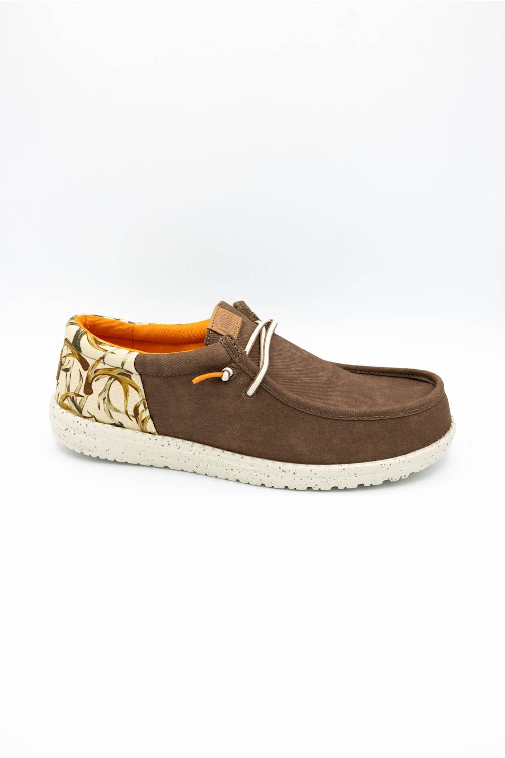 A brown suede slip-on shoe with a patterned heel panel, white laces, and a white speckled sole, shown against a plain white background.
