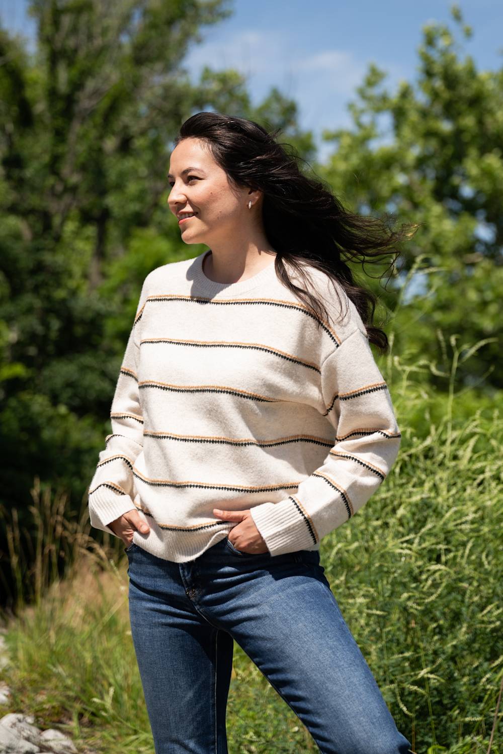 A model stands outdoors wearing a cream knit sweater with thin black and tan horizontal stripes, paired with dark blue jeans. Green trees and tall grass fill the background on a sunny day.