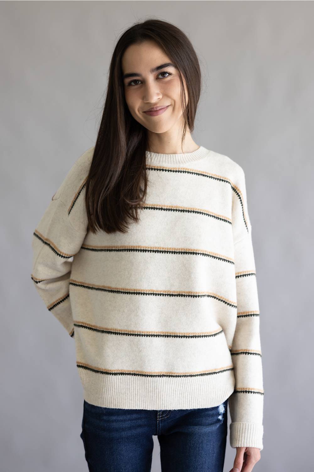 A woman wearing a cream-colored knit sweater with thin horizontal black and tan stripes, paired with dark blue jeans, standing against a neutral gray background.
