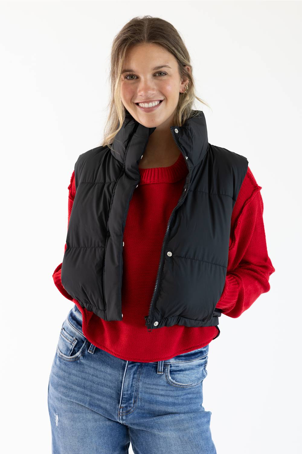 A woman wearing a black quilted puffer vest over a red long-sleeve top and blue jeans, photographed against a plain white studio background.