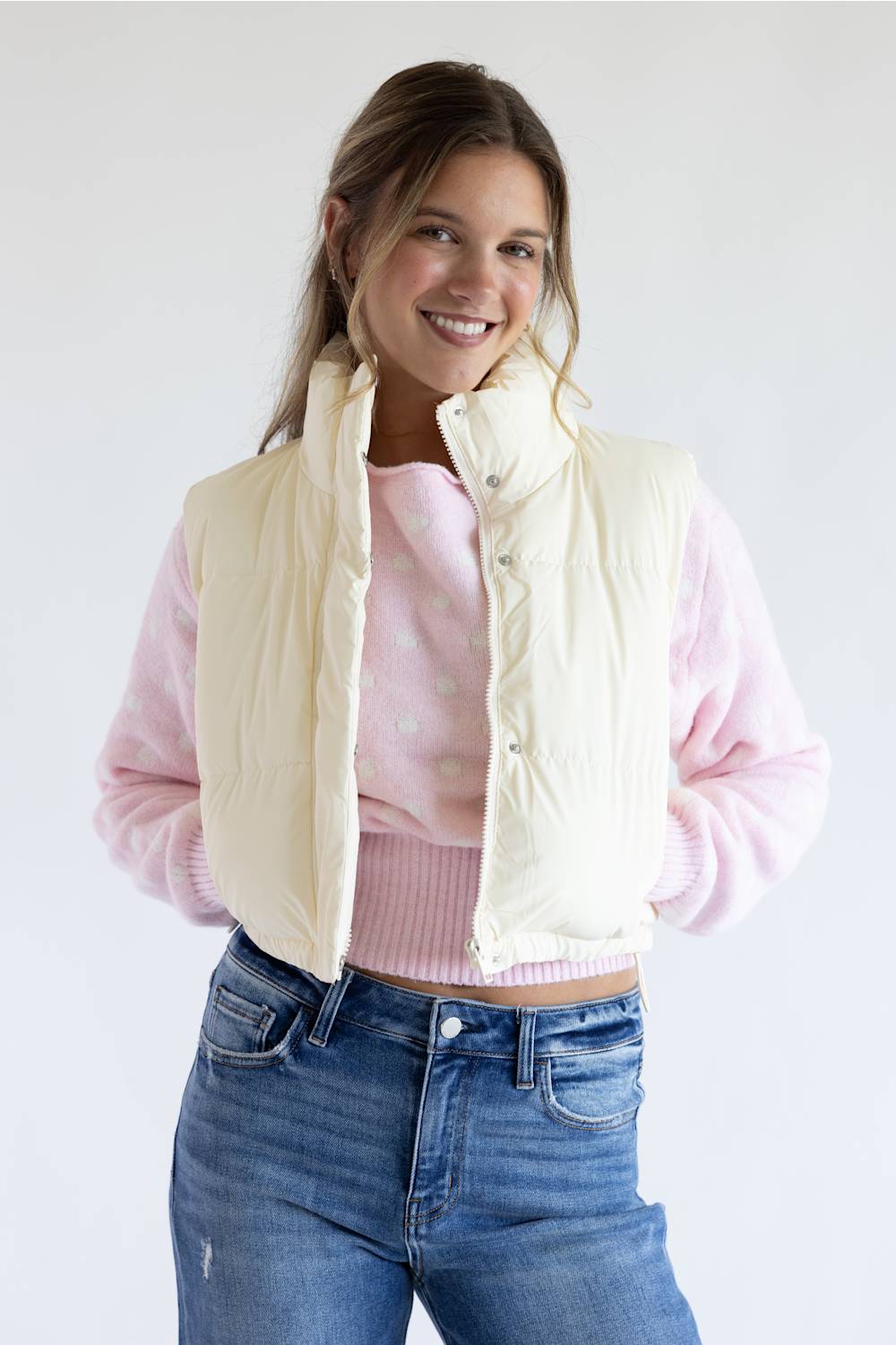 A woman modeling a cream-colored sleeveless puffer vest over a pink long-sleeve top and blue jeans, photographed against a plain light gray studio background.