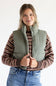A woman wearing an olive green puffer vest over a brown and beige striped knit sweater, paired with blue jeans, against a plain white studio background.