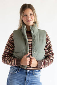 A woman wearing an olive green puffer vest over a brown and beige striped knit sweater, paired with blue jeans, against a plain white studio background.