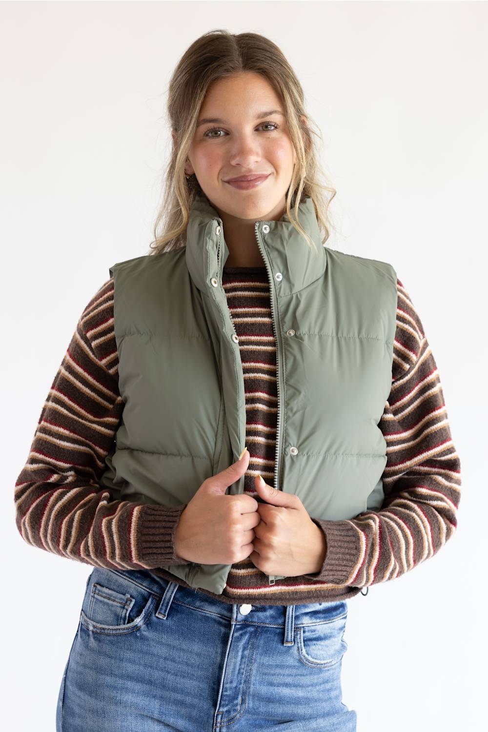 A woman wearing an olive green puffer vest over a brown and beige striped knit sweater, paired with blue jeans, against a plain white studio background.