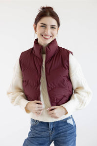 A smiling woman wearing a burgundy quilted puffer vest over a cream cable-knit sweater and blue jeans, photographed against a plain white studio background.