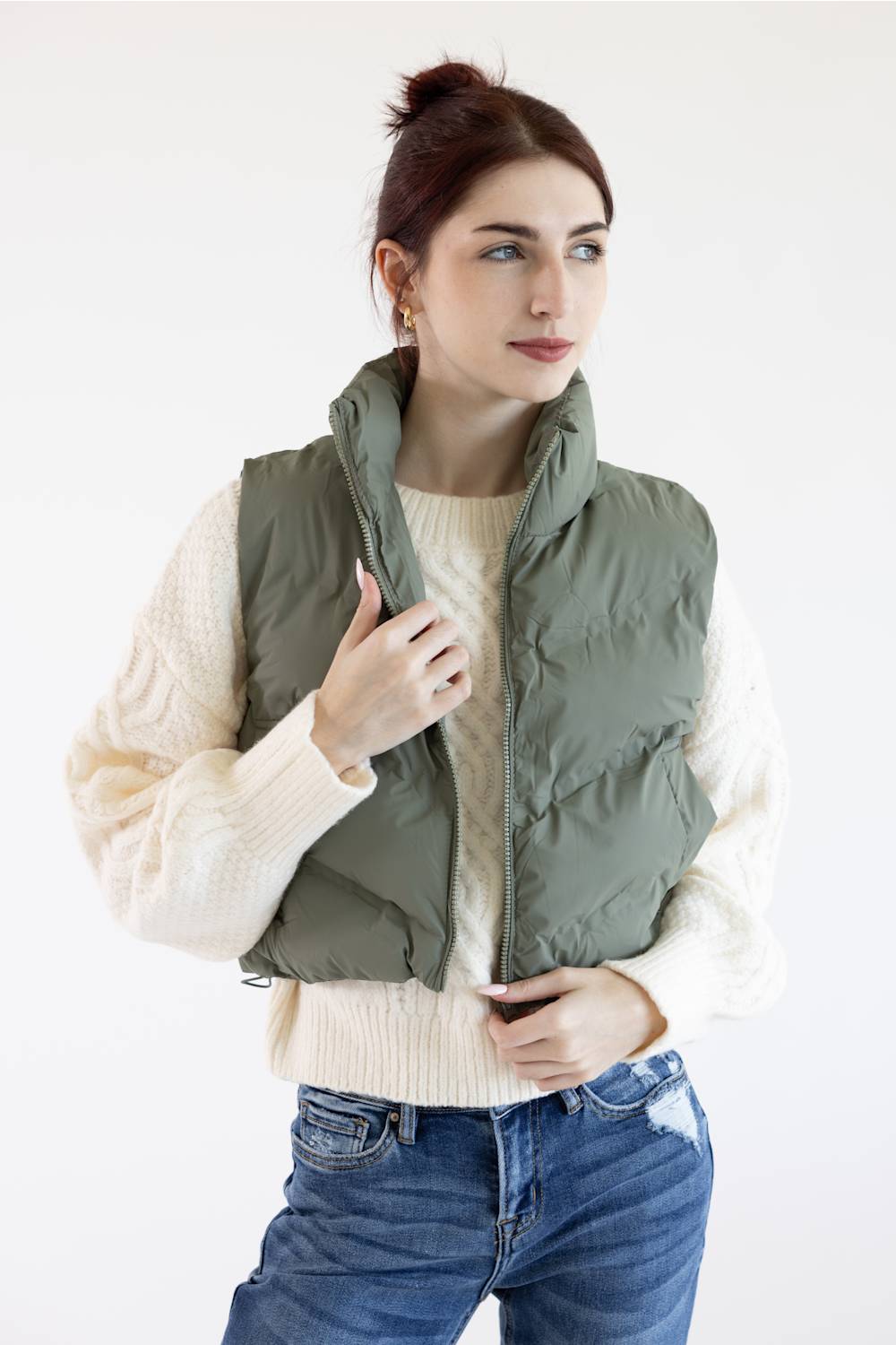 A woman wearing an olive green puffer vest over a cream cable-knit sweater and blue jeans, standing against a plain white studio background.