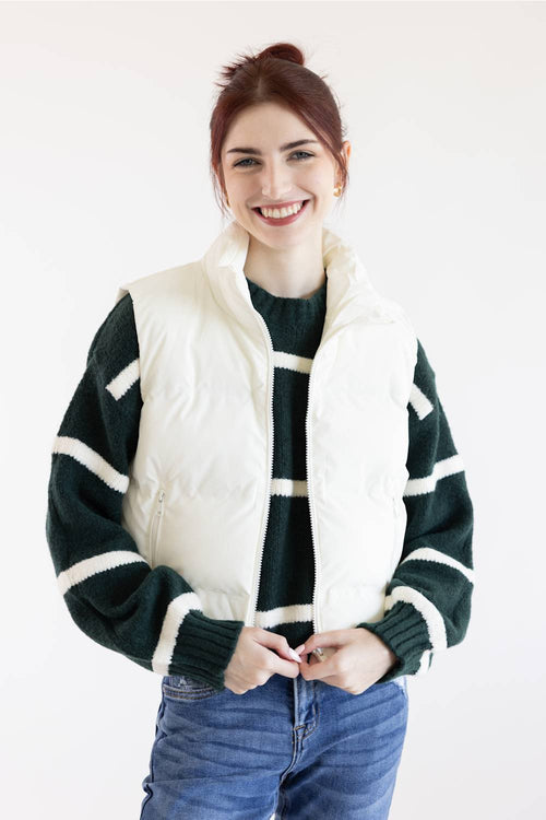 A smiling woman wearing a cream quilted puffer vest over a dark green and cream striped knit sweater, paired with blue jeans, against a plain white studio background.