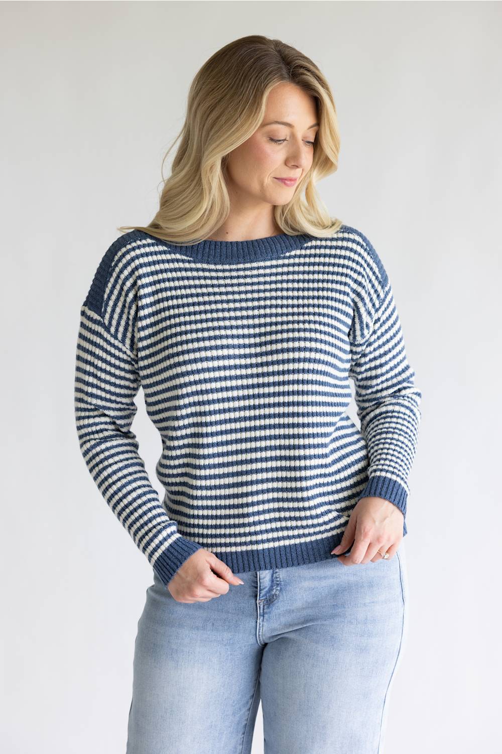A woman wearing a navy and white horizontally striped knit sweater with a boat neckline, paired with light blue jeans, standing against a plain light gray background.