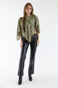 A woman modeling a sage green patterned poncho top paired with black leather flare pants and black ankle boots, photographed against a seamless white studio background.