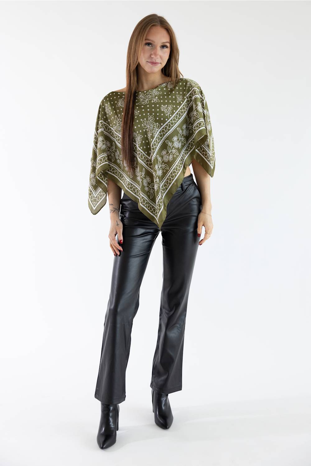 A woman modeling a sage green patterned poncho top paired with black leather flare pants and black ankle boots, photographed against a seamless white studio background.