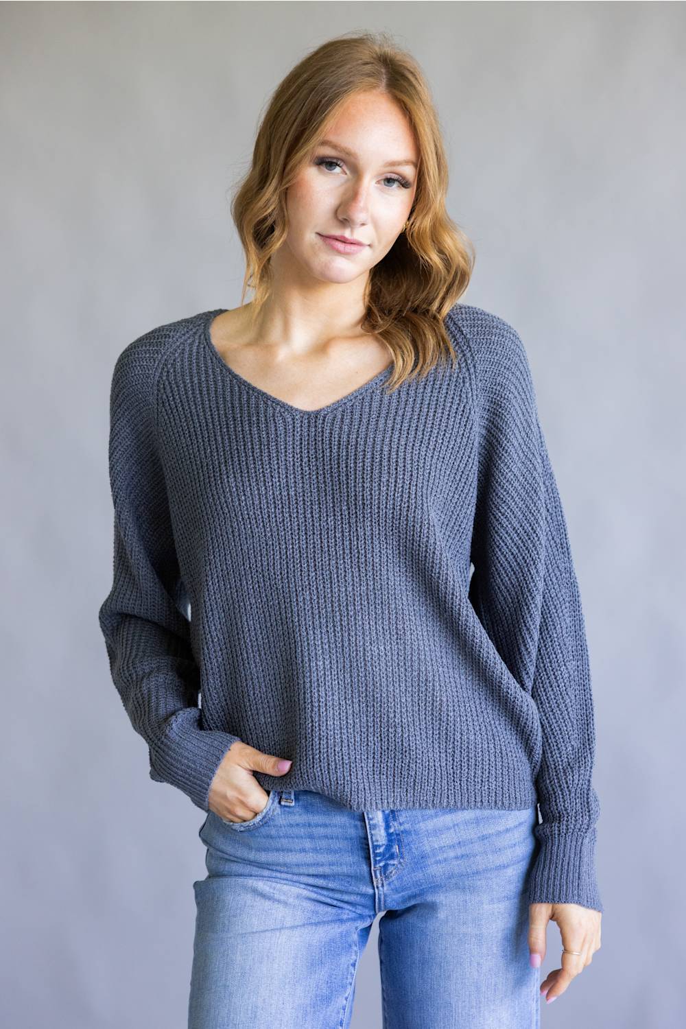 A woman wearing a slate blue ribbed knit sweater with a V-neck and long sleeves, paired with blue jeans, standing against a neutral gray studio background.
