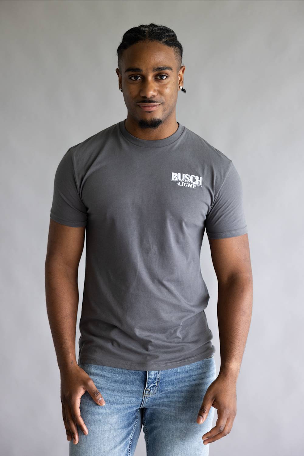 A man wearing a dark gray crewneck t-shirt with a white 'BUSCH LIGHT' logo on the left chest, paired with light blue jeans, standing against a neutral gray studio background.