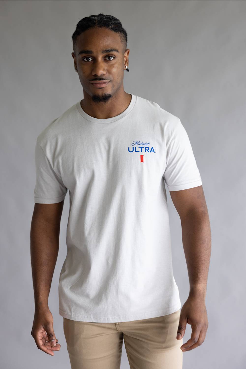 A man wearing a white short-sleeve crewneck t-shirt with a blue and red 'Michael ULTRA' logo on the left chest, paired with beige pants, against a neutral gray studio background.