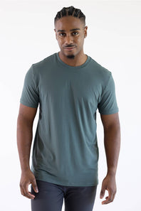 A man with braided hair wearing a dark green crewneck t-shirt and grey pants, standing against a plain white background.