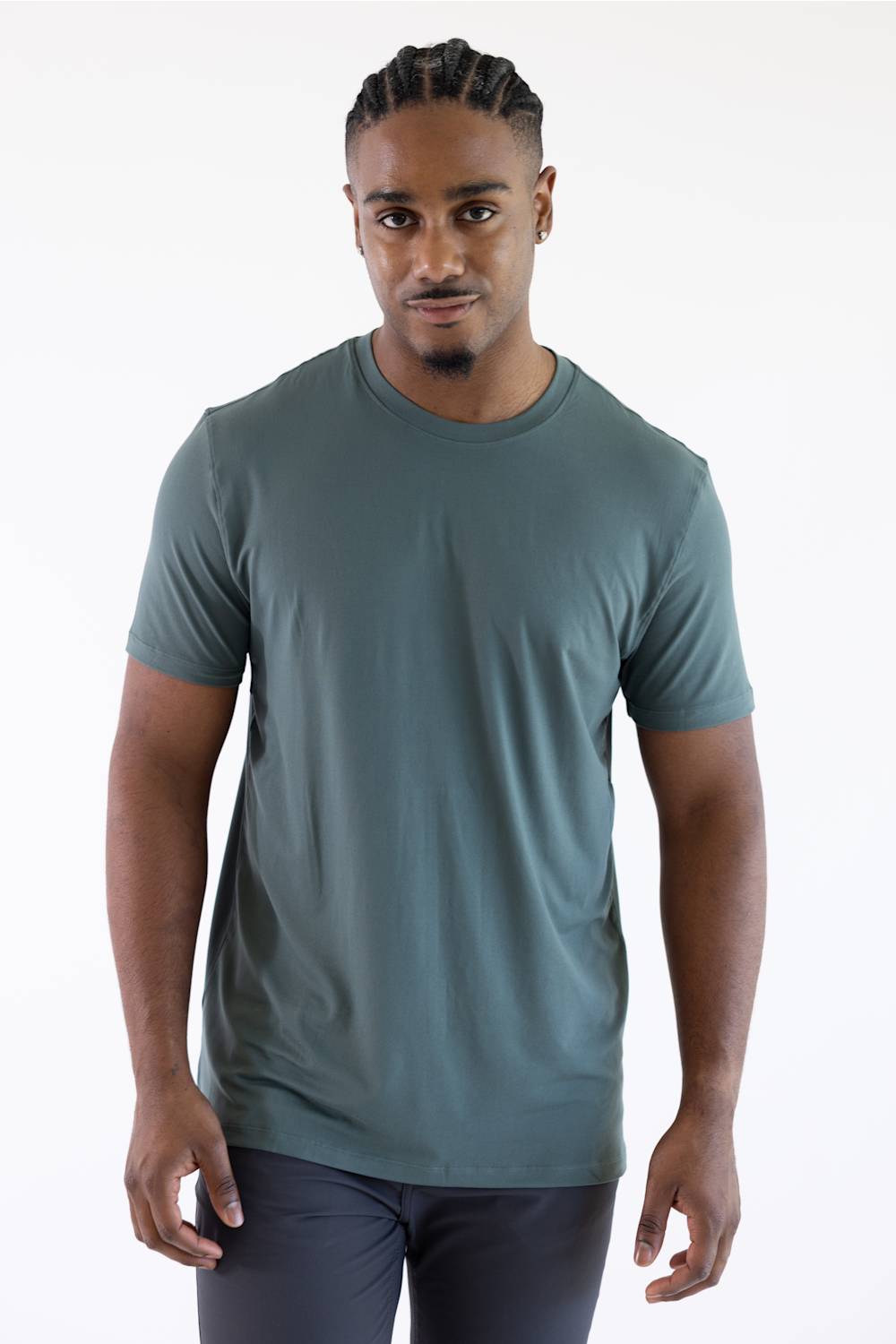 A man with braided hair wearing a dark green crewneck t-shirt and grey pants, standing against a plain white background.