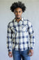 A man wearing a long-sleeve plaid button-up shirt in white, navy, and gray, paired with blue jeans, standing against a plain light gray background.