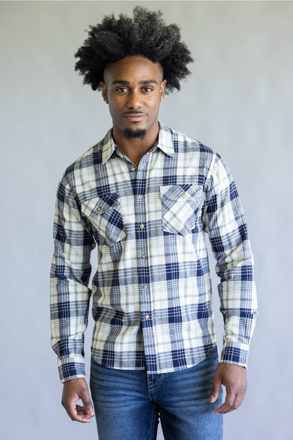 A man wearing a long-sleeve plaid button-up shirt in white, navy, and gray, paired with blue jeans, standing against a plain light gray background.