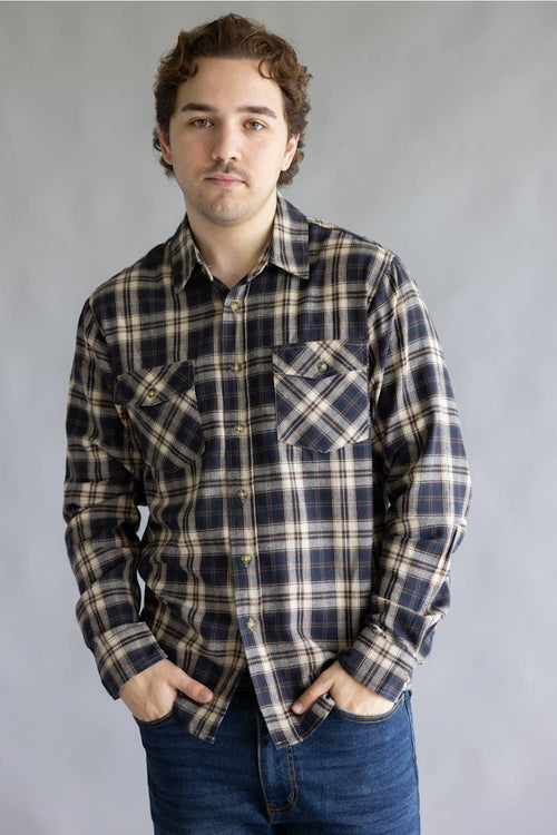A man wearing a long-sleeve plaid button-up shirt in brown, beige, and white, paired with blue jeans, standing against a neutral gray studio background.