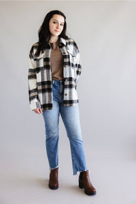 A woman modeling a black, white, and brown plaid flannel shirt jacket over a brown top, paired with blue jeans and brown boots, against a plain light gray studio background.