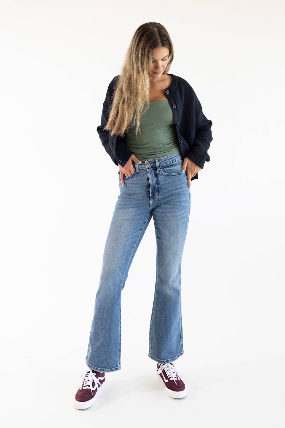 A woman modeling a pair of light-wash blue flare jeans, styled with a green tank top, a dark navy cardigan, and burgundy sneakers, against a plain white studio background.