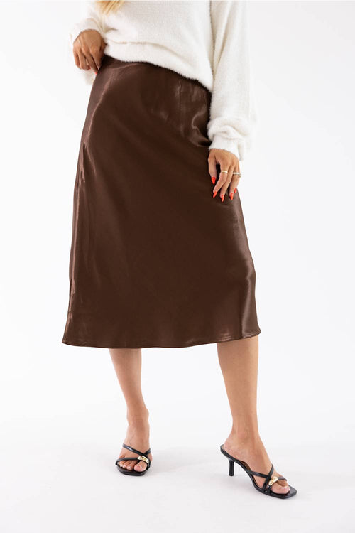 A model wearing a brown midi skirt with a satin-like sheen, styled with a white sweater and black heeled sandals against a white studio background.