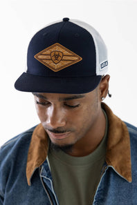 A man wearing a navy blue and white trucker hat with a brown diamond-shaped patch on the front, styled with a denim jacket and olive green t-shirt against a plain white background.
