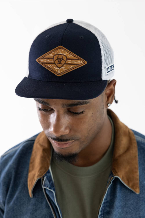 A man wearing a navy blue and white trucker hat with a brown diamond-shaped patch on the front, styled with a denim jacket and olive green t-shirt against a plain white background.