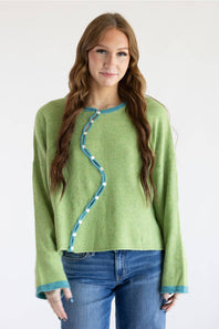 A woman modeling a light green knit sweater with a distinctive wavy blue trim and white button details down the front, paired with blue jeans against a plain white background.