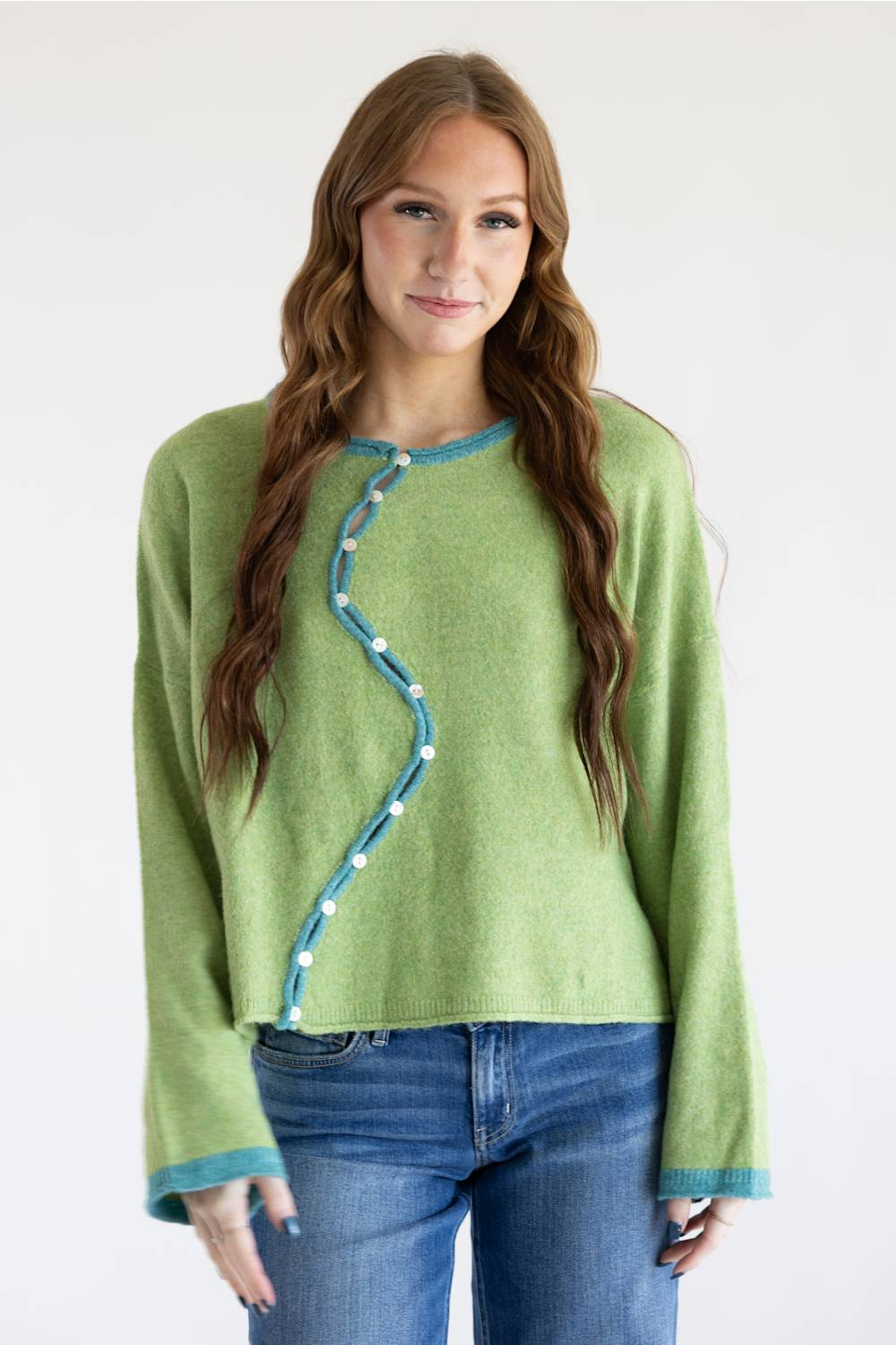 A woman modeling a light green knit sweater with a distinctive wavy blue trim and white button details down the front, paired with blue jeans against a plain white background.