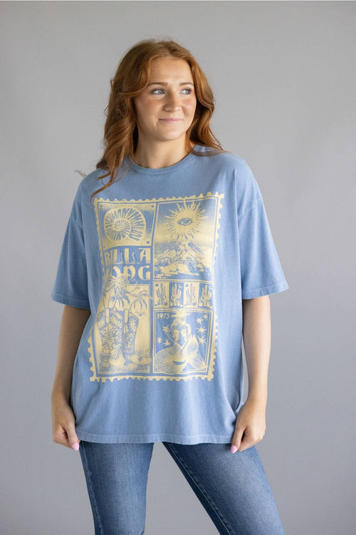 A woman modeling a light blue oversized graphic t-shirt with a large yellow vintage-style print on the front, paired with blue jeans, against a solid gray studio background.