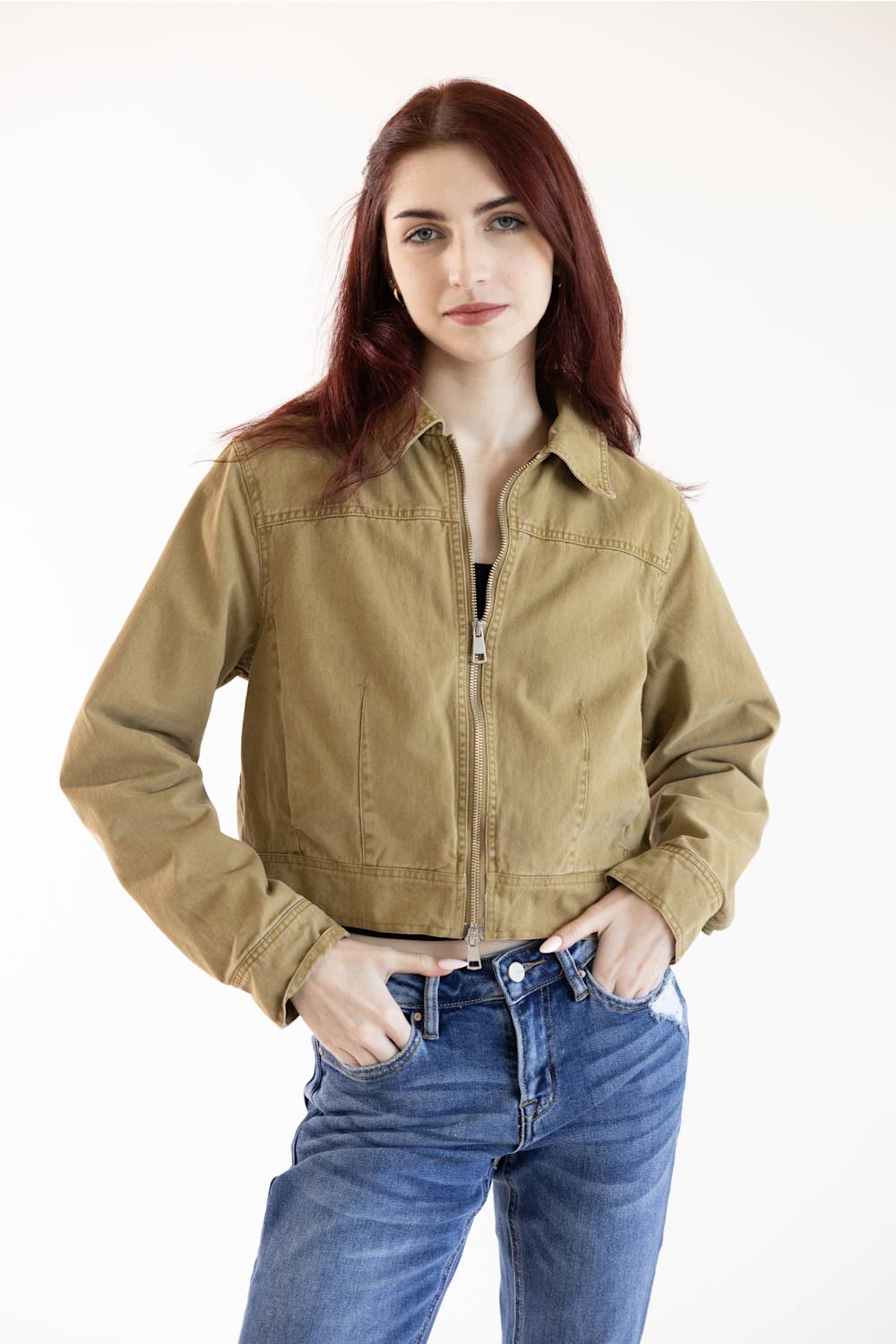 A woman with red hair wearing a cropped tan zip-up jacket and blue jeans, standing against a plain white studio background.