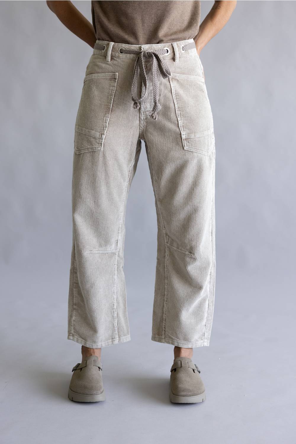 A person wearing light beige wide-leg cropped pants with a drawstring waist, paired with a taupe top and taupe slip-on shoes, shown against a neutral gray studio background.