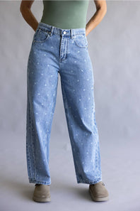 A person wearing light blue high-waisted, wide-leg denim jeans with a subtle white speckled pattern, styled with a sage green top and beige slip-on shoes against a neutral gray studio background.