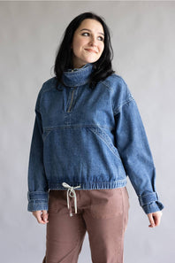 A woman wearing a blue denim half-zip pullover sweater with a high mock neck and brown pants, standing against a neutral gray studio background.