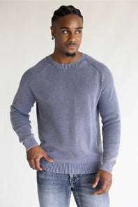 A man wearing a ribbed knit crewneck sweater in a dusty periwinkle blue color, paired with blue jeans, standing against a plain off-white background.