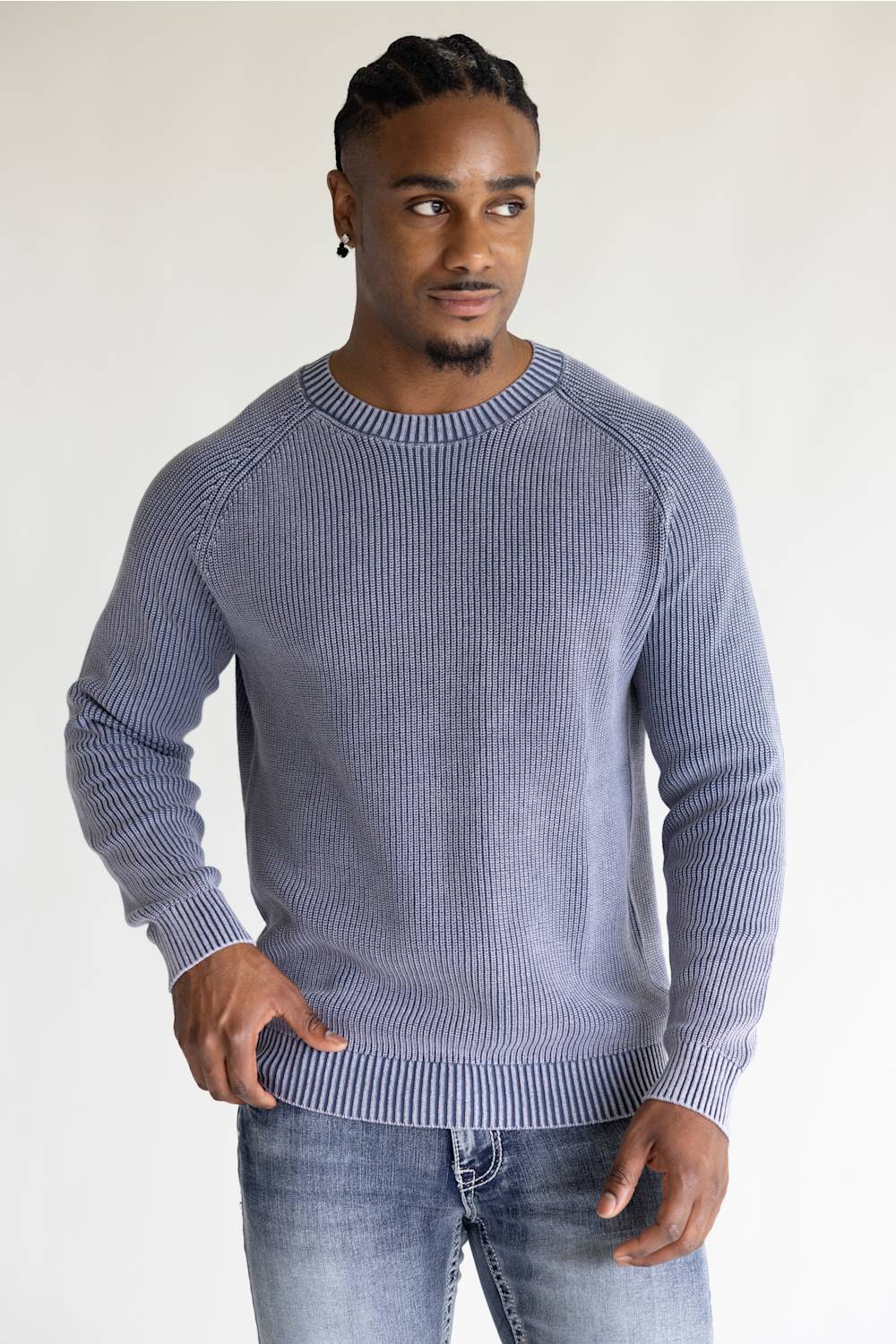 A man wearing a ribbed knit crewneck sweater in a dusty periwinkle blue color, paired with blue jeans, standing against a plain off-white background.
